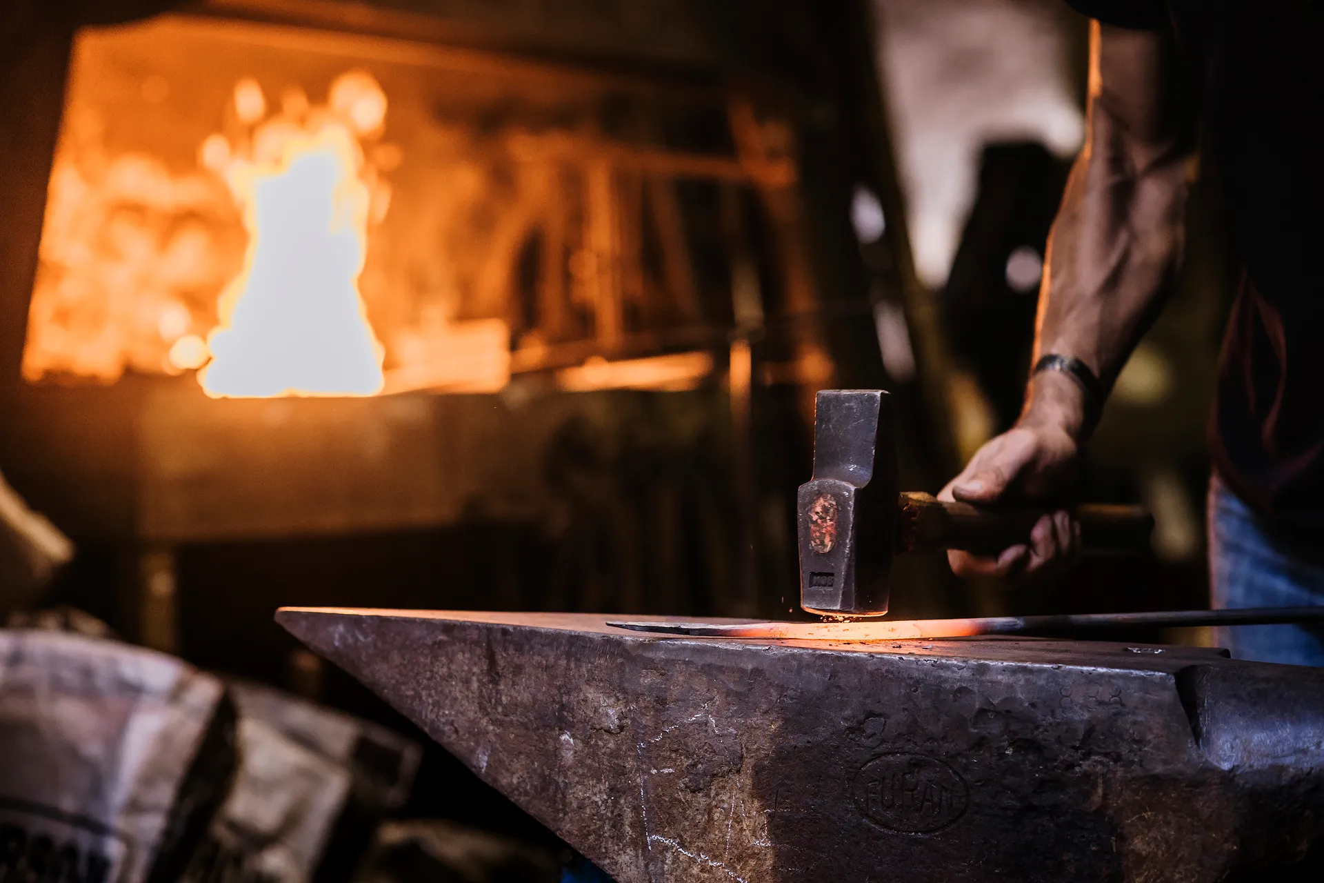 un bras musclé frappe du marteau sur une barre de fer rougie, un feu à l'arrière-plan