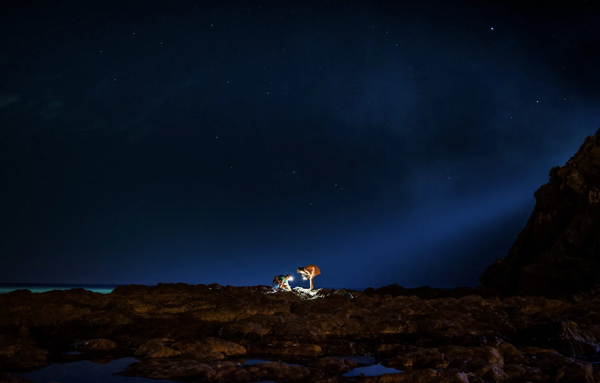 Deux personnes s'éclairent à la lampe sur un paysage de rochers au bord de la mer, paysage nocturne
